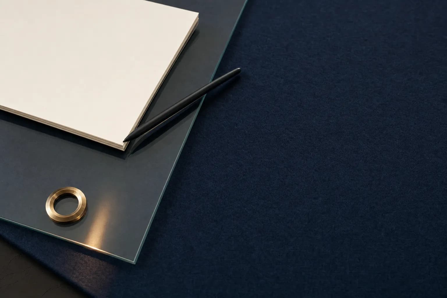 A slim black stylus resting on a sheet of glass over dark navy felt beside a stack of blank cream paper and a brass ring