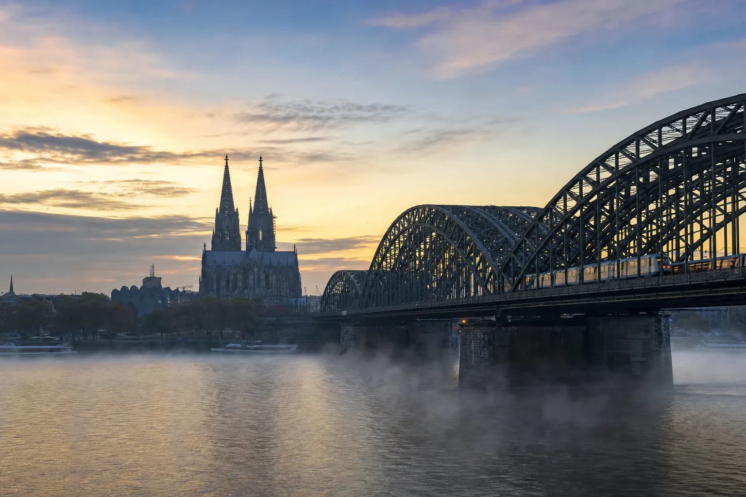 Cologne Cathedral and the Hohenzollern railway bridge over the Rhine at dawn, mist on the water and a train crossing