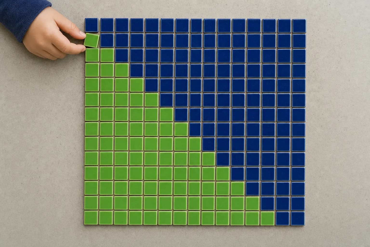An overhead view of a square grid of small ceramic tiles on a pale table, navy above the diagonal and green below it, with a child's fingertip lifting one corner tile