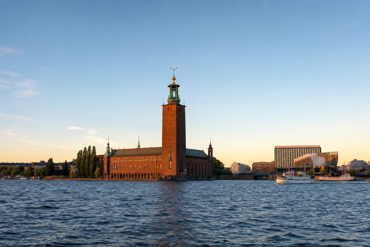 Stockholm City Hall and its brick tower seen across the water of Riddarfjärden in golden evening light, with two white ferries passing